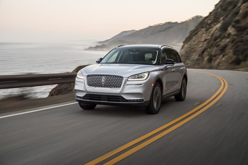 Silver Lincoln Corsair SUV driving on a winding coastal highway next to the ocean under a soft hazy sky.