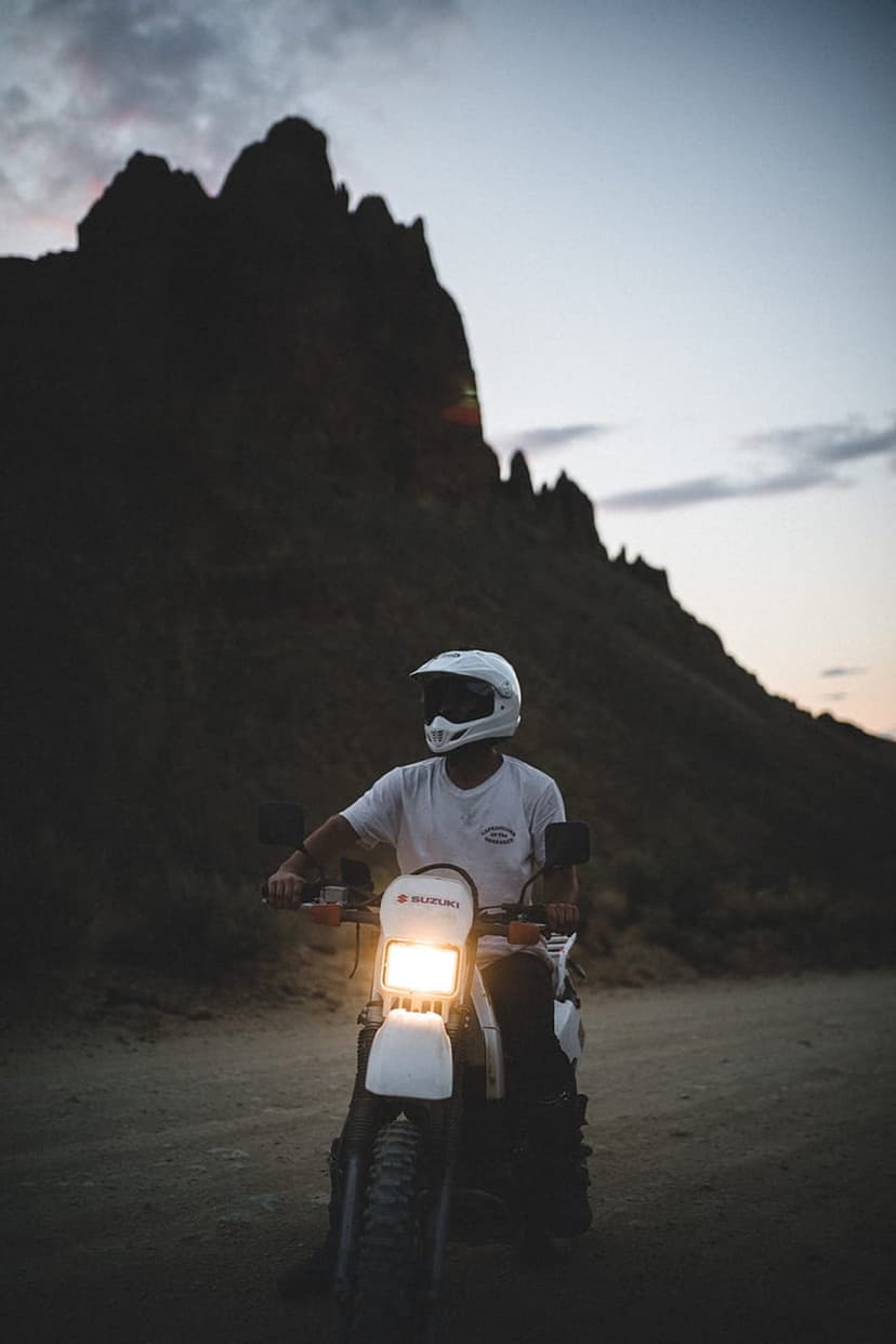 Motorcyclist in a white helmet sits on a white dirt bike with its headlight on before dark mountain silhouettes.