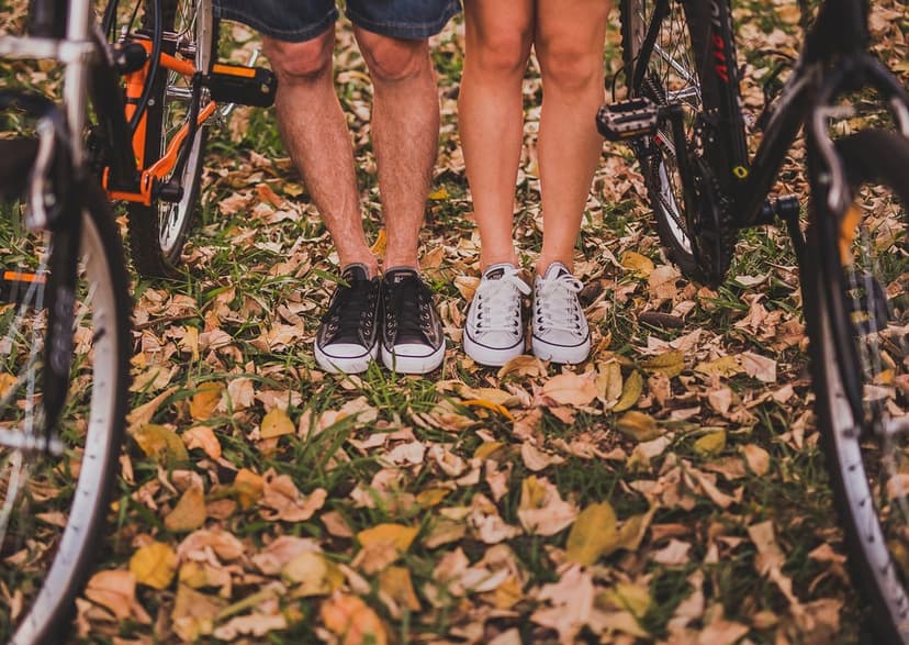 Couple’s feet in black and white sneakers standing on fallen orange leaves between two bicycle wheels outdoors.