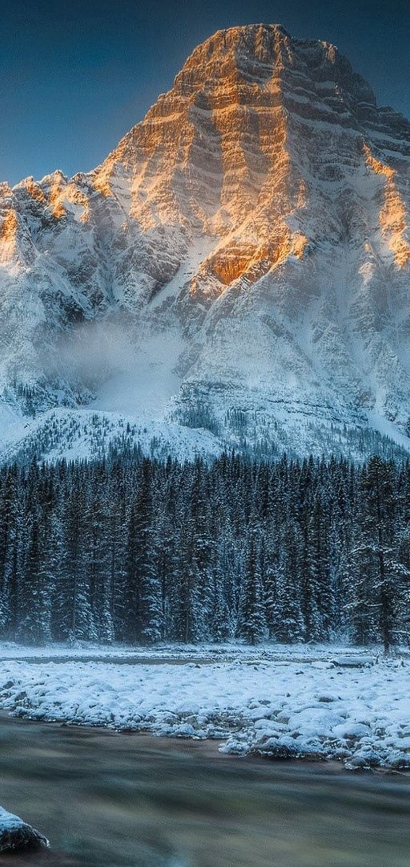 Snow-capped mountain peak glowing with golden sunlight above a frozen river and evergreen forest in winter.