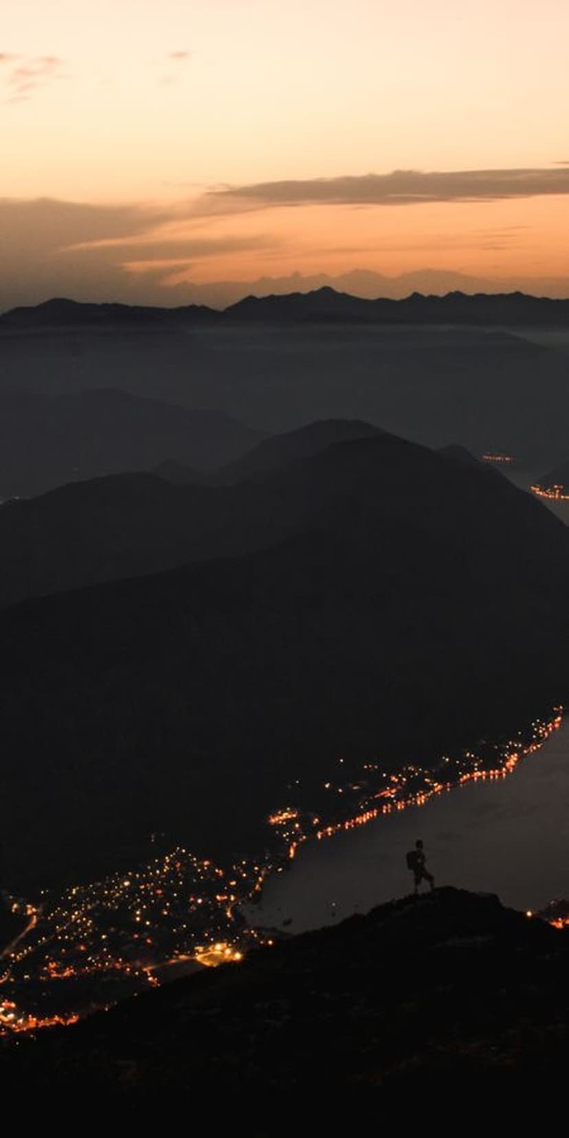 Solitary person stands on a high cliff overlooking a glowing lakeside town nestled between dark mountain peaks.