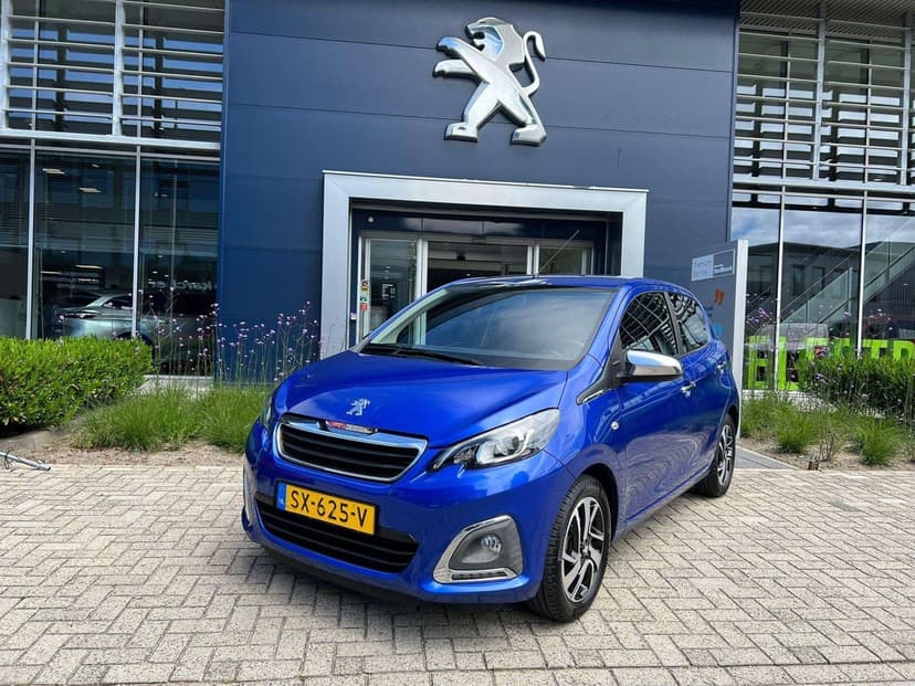 Blue Peugeot 108 hatchback parked on a paved driveway in front of a dealership with a large silver lion logo.