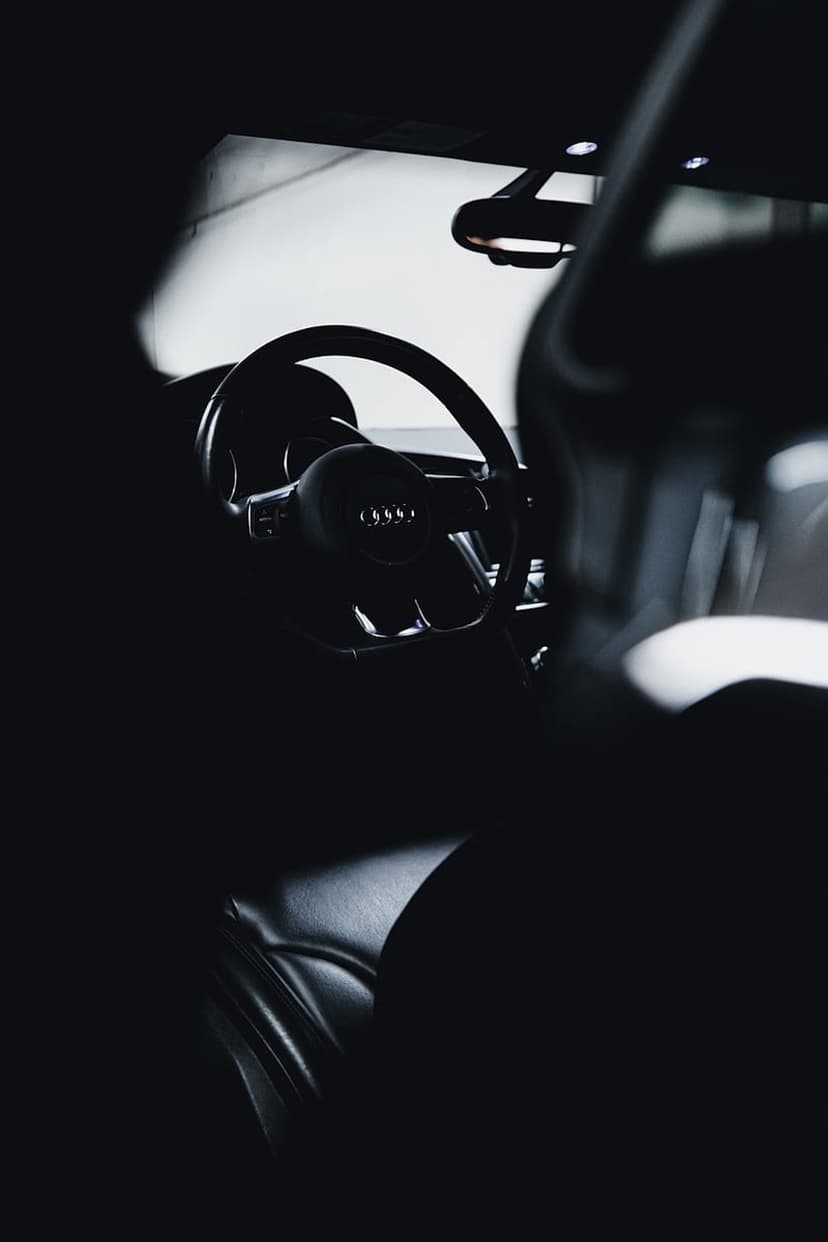 Audi steering wheel logo centered in a dark leather car interior with shadowed driver seats and premium trim.