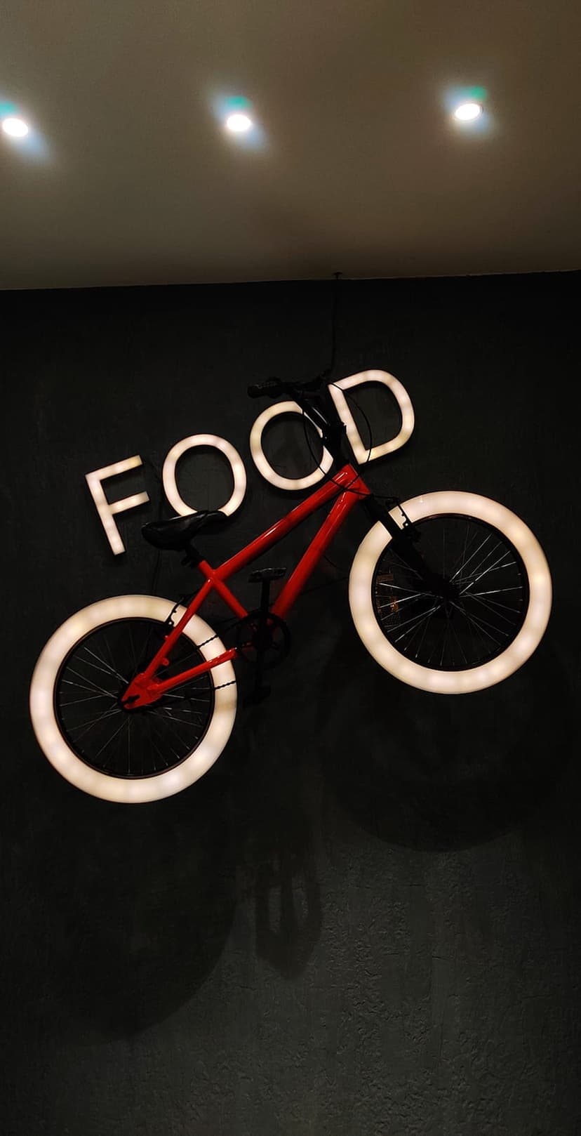 Red bicycle mounted on a dark wall with white neon circles on the wheels and a glowing Food sign above it.