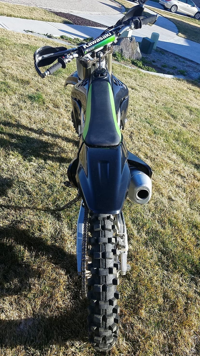 Black and green Kawasaki dirt bike with knobby tires parked on a grassy slope showing the rear fender and exhaust.