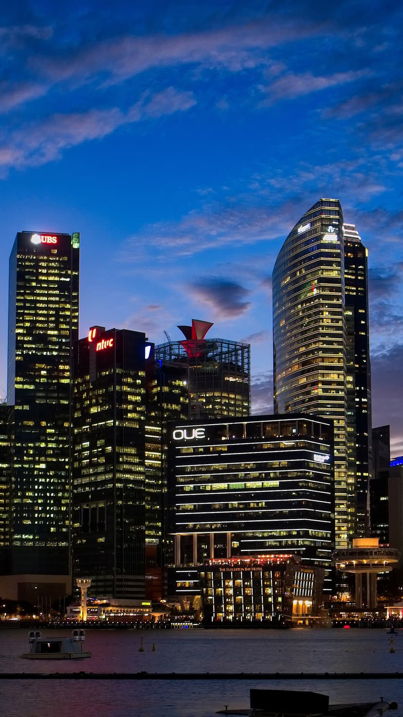 Singapore skyscrapers glow with golden lights against a dark twilight sky, reflecting in the calm bay waters.
