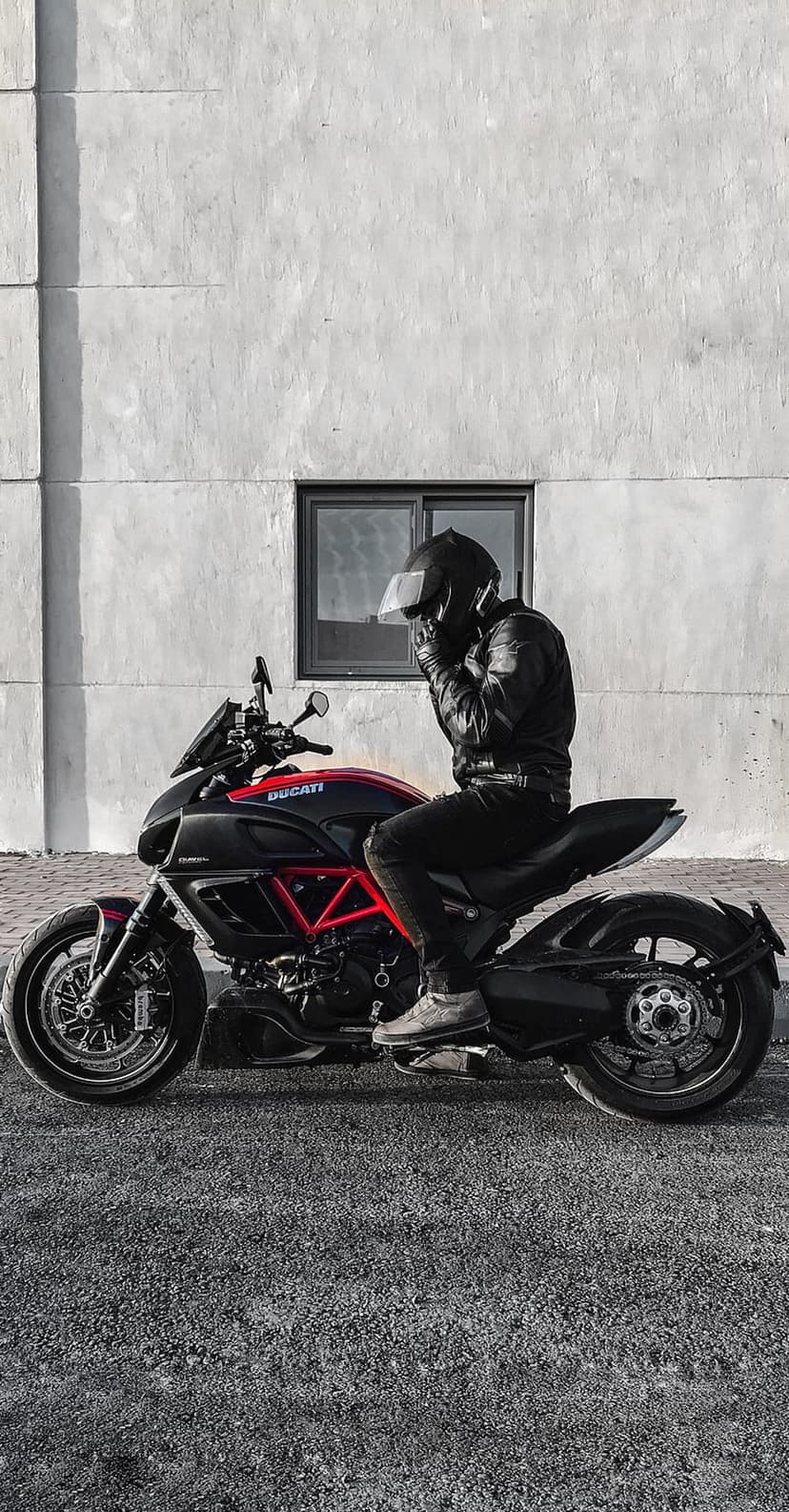 Rider wearing a black leather jacket and helmet sits on a red and black Ducati motorcycle against a white wall.
