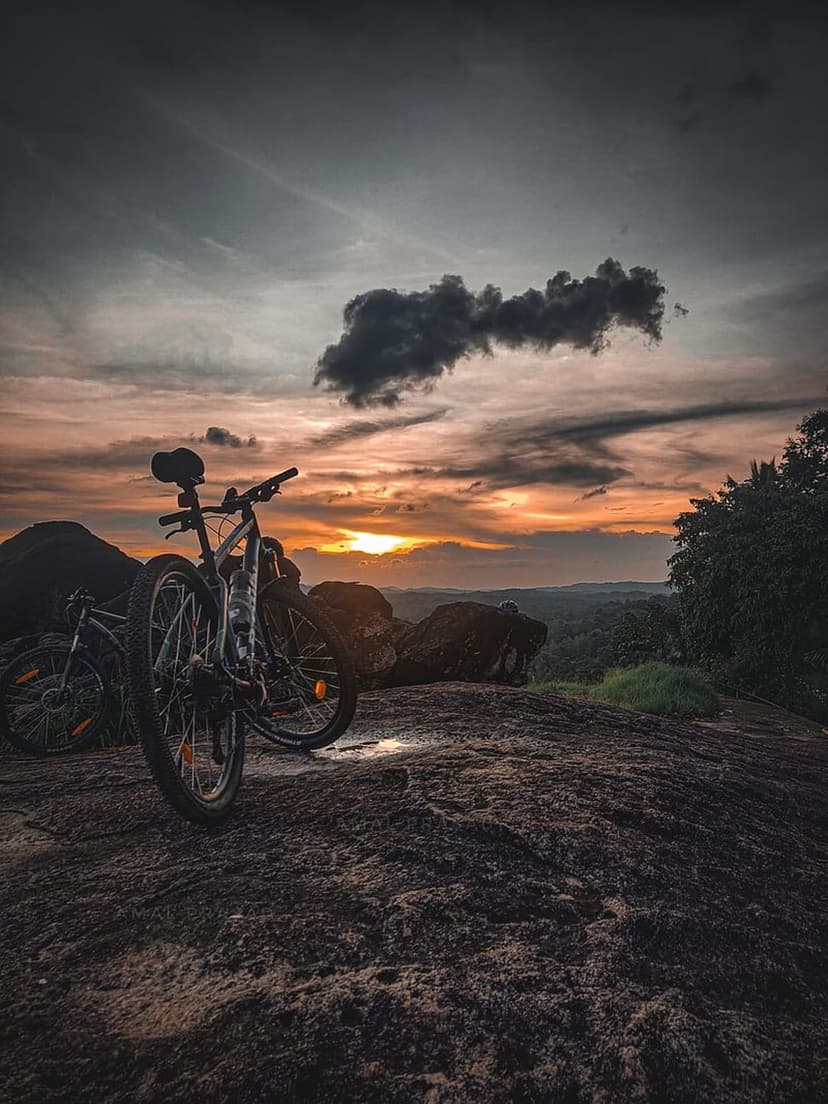 Two mountain bikes on a jagged rock formation under a dark swirling sunset sky overlooking a distant green valley.