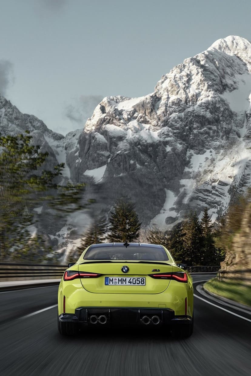 Rear view of a yellow BMW M4 sports car driving on a mountain road with snow-covered peaks and blurred scenery.