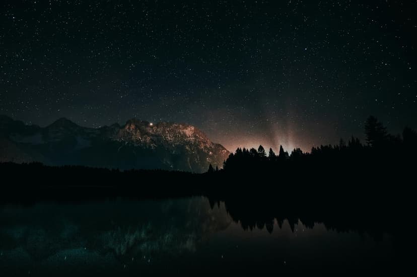 Jagged mountain peaks under a dense field of stars reflected in a still lake with a dark forest silhouette.