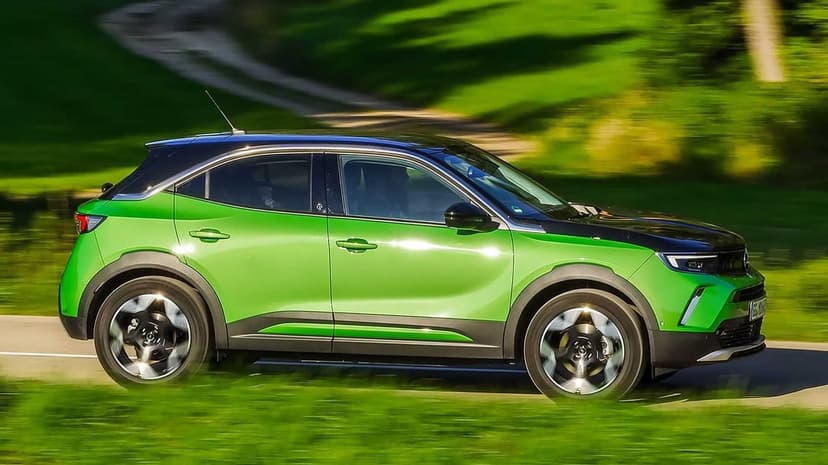 Green and black electric SUV driving on a road with motion blur against a sunny green hill background.