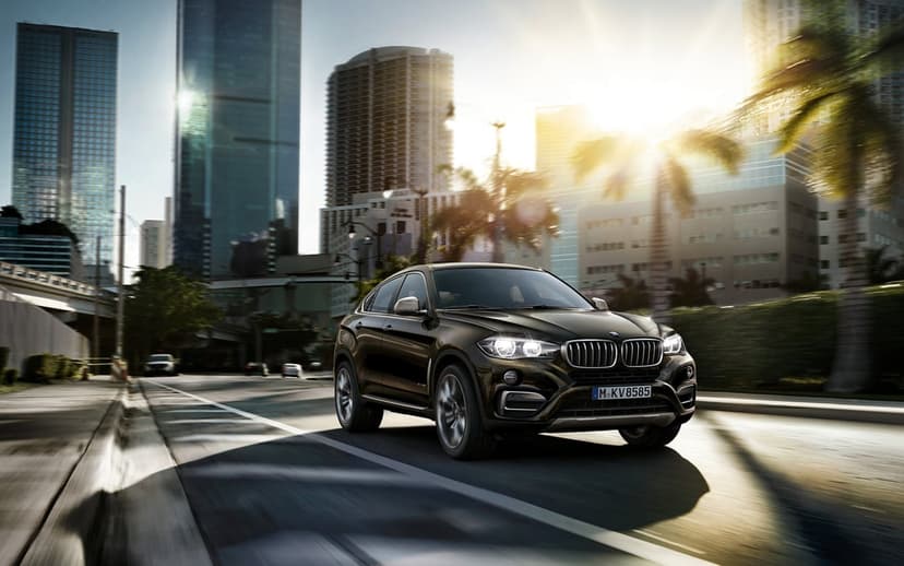Dark brown BMW X6 driving on an urban street with motion blur, skyscrapers, and golden sunset lighting.