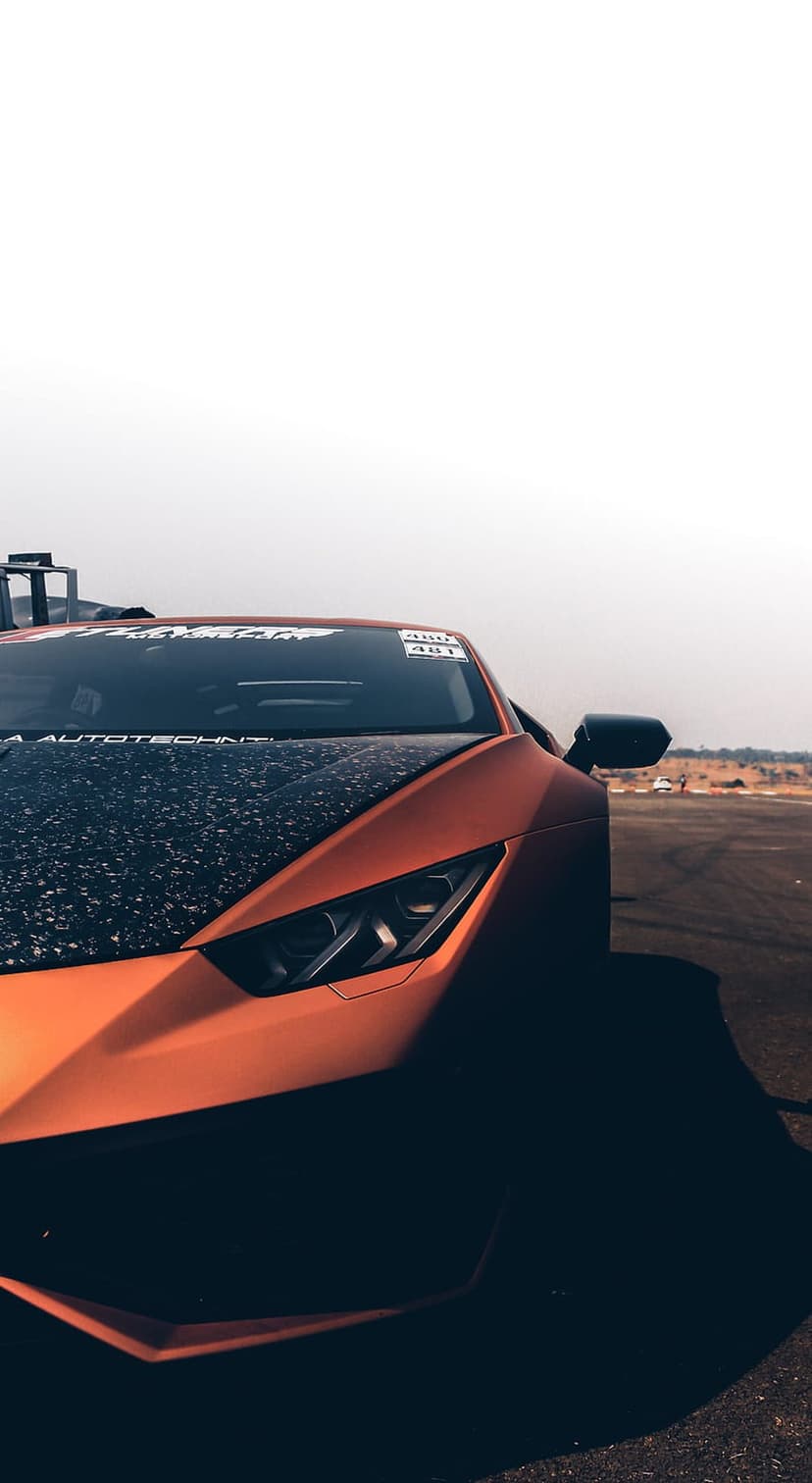 Matte orange Lamborghini with a carbon fiber hood and lit headlights parked on an asphalt track under a hazy sky.