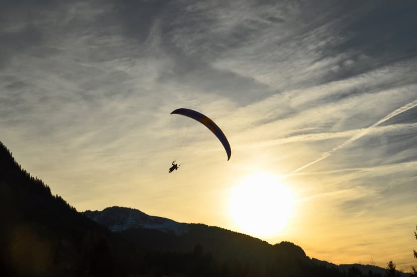 Paraglider silhouette floating above jagged mountain peaks during a radiant golden hour sunset with glowing clouds.