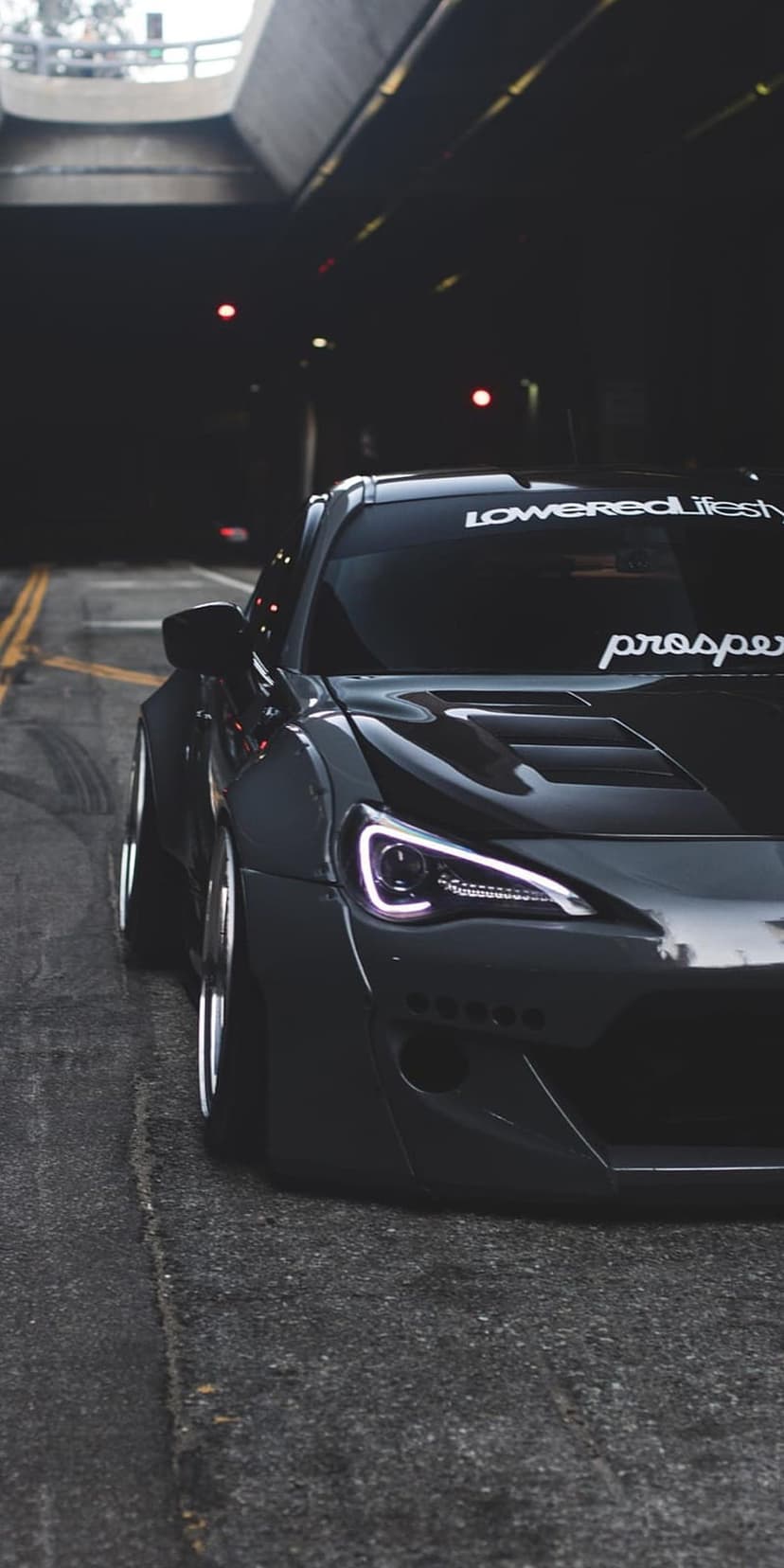 Slammed black sports car with purple headlights parked on wet asphalt beneath a concrete bridge at night.
