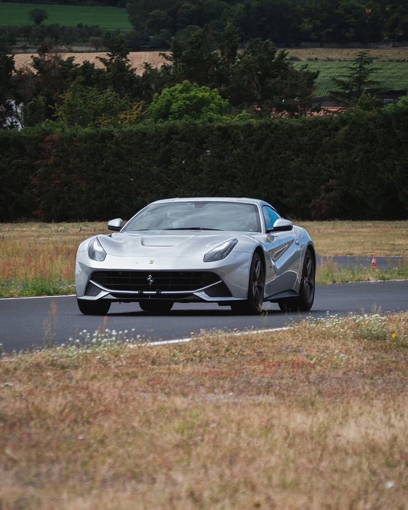 Silver Ferrari sports car speeding on a paved track surrounded by green trees and dry grass hills.