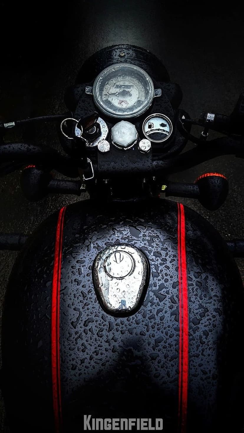 Raindrops on a black motorcycle fuel tank featuring red stripes, a chrome speedometer, and fuel cap.