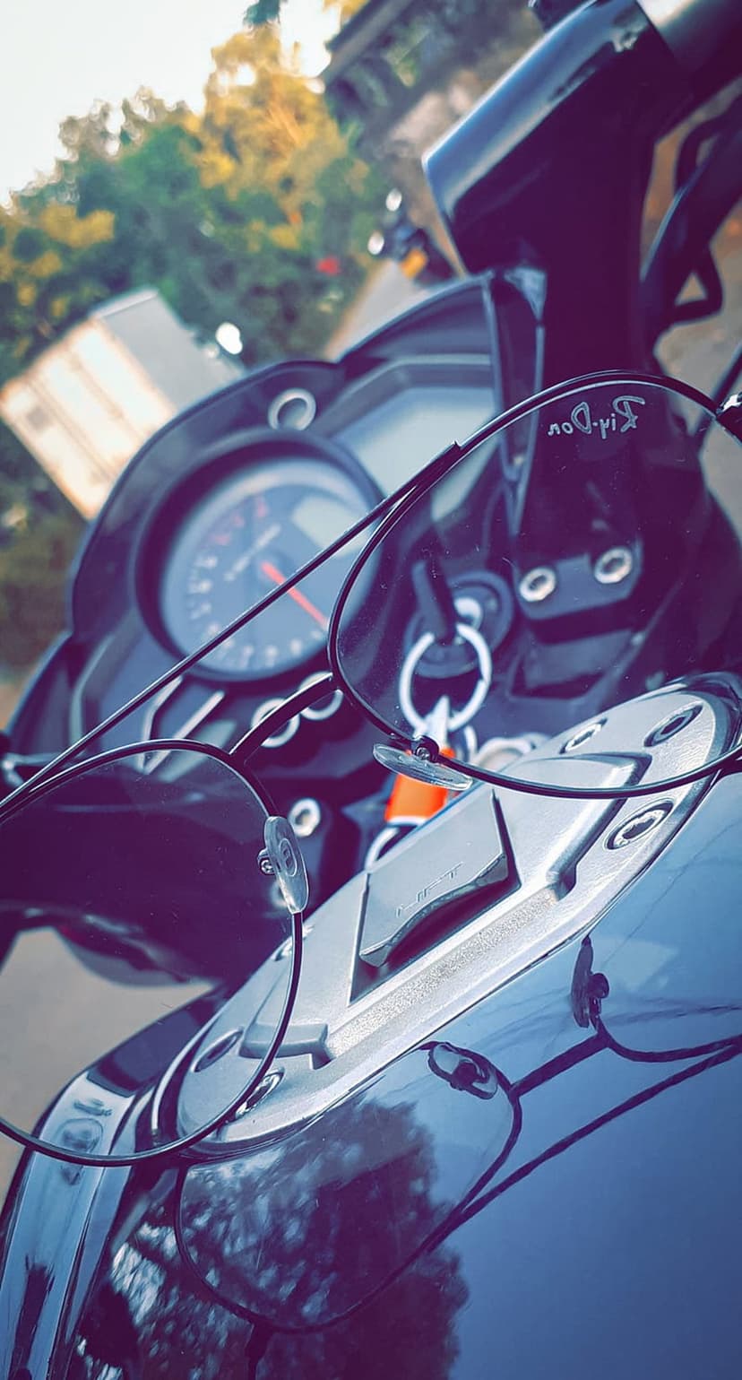 Aviator sunglasses resting on a chrome motorcycle dashboard reflect a blue sky and green trees in their lenses.