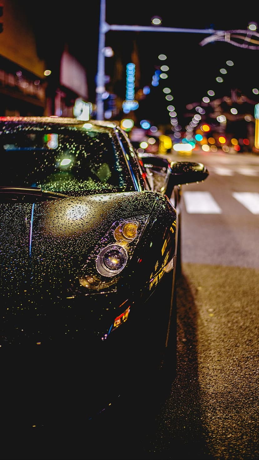 Black luxury car parked on wet asphalt with rain droplets reflecting colorful city bokeh during a stormy night.