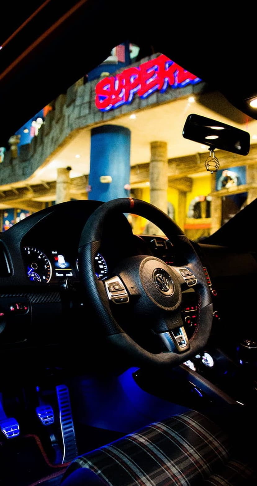 Custom car interior glows with blue ambient light across a sporty steering wheel, plaid seat, and metal pedals.