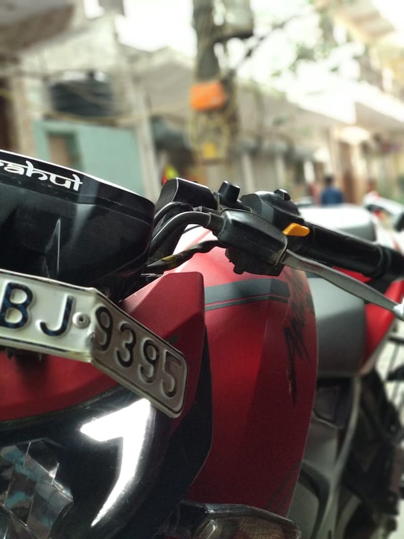 Red motorcycle front assembly with chrome handlebars and BJ 9395 license plate against a blurred background.