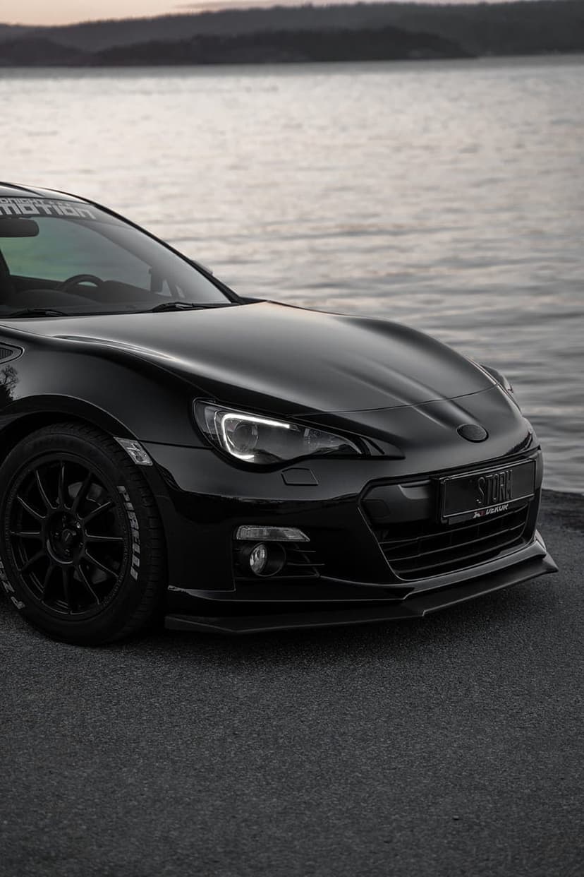 Black Subaru BRZ with LED headlights and a front lip spoiler parked on a paved surface next to calm water.