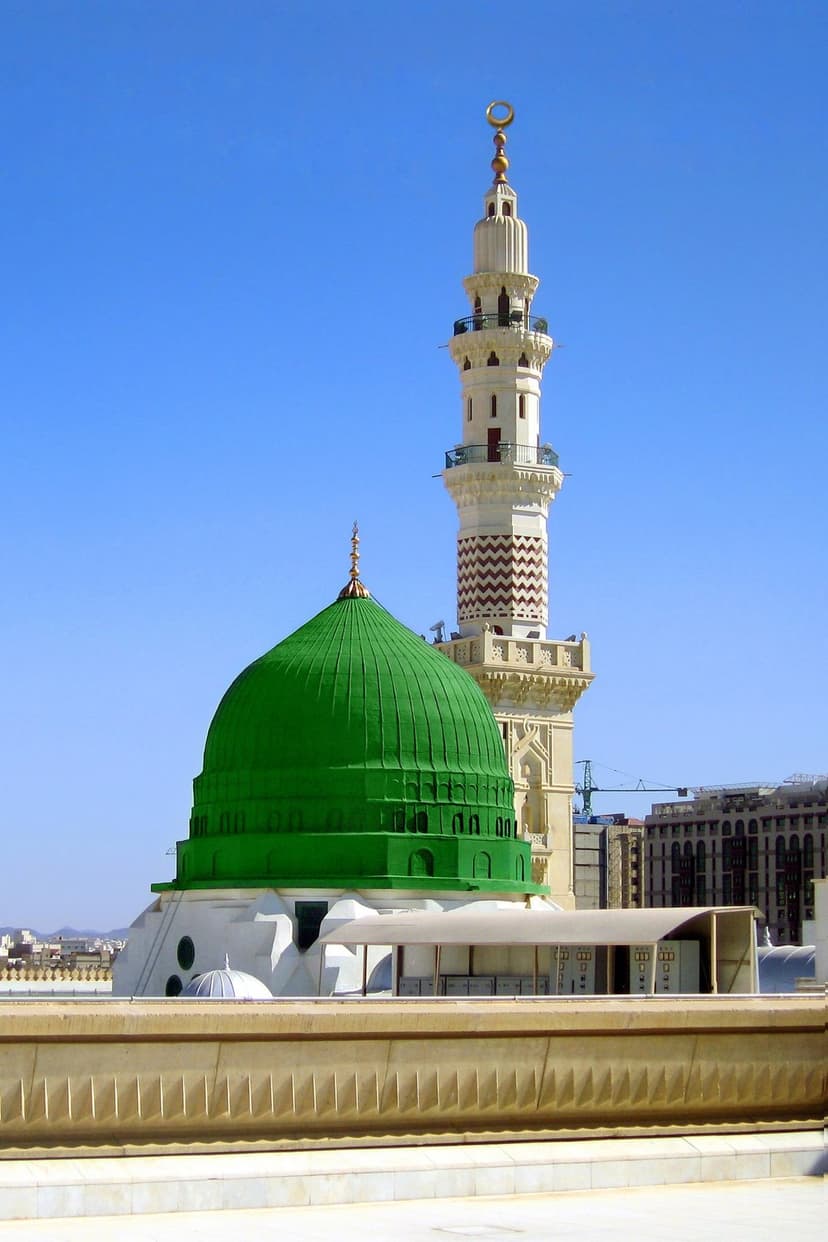 Green dome and a towering stone minaret of the Prophet's Mosque in Medina stand under a clear blue morning sky.