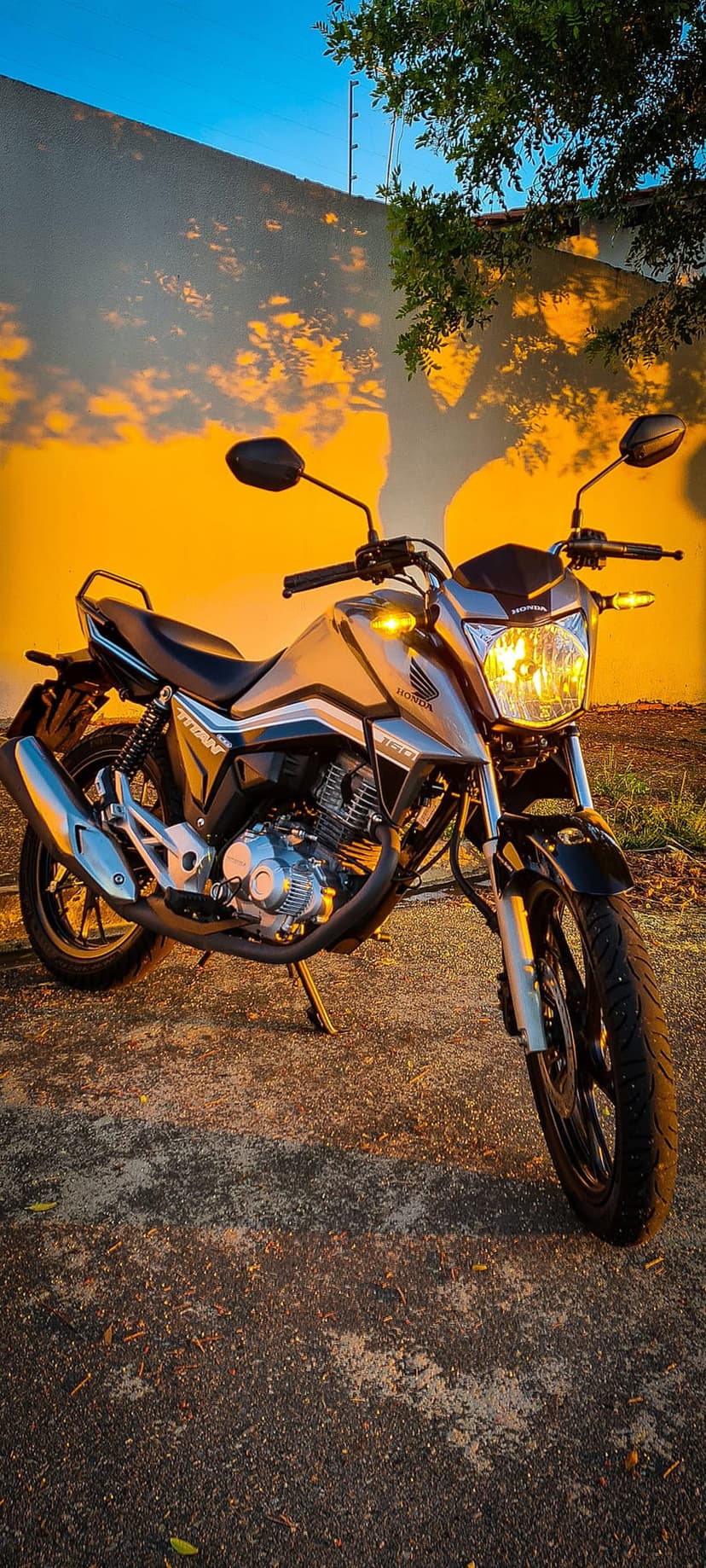 Honda Titan motorcycle parked outdoors in golden hour light with tree shadows against a vivid orange wall.