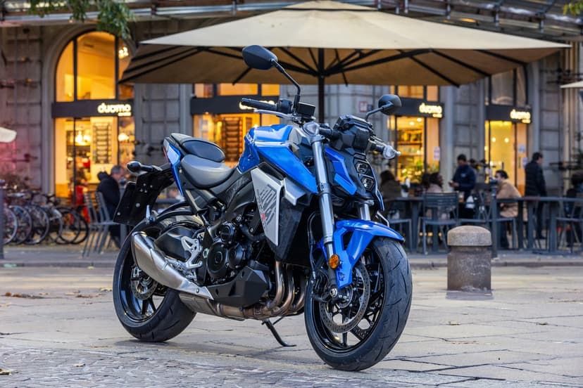 Blue Suzuki GSX-S1000 motorcycle parked on cobblestones before a blurred urban cafe with a large beige umbrella