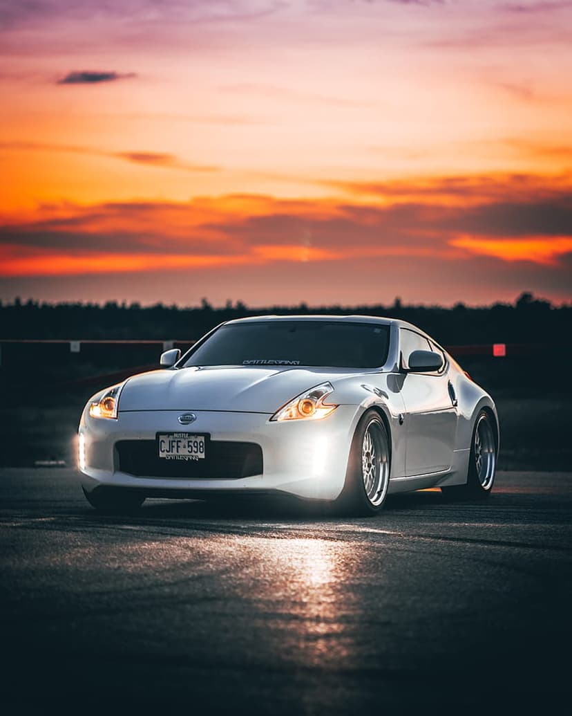 White Nissan 370Z sports car parked on asphalt under a vivid orange and purple sunset sky with glowing lights.