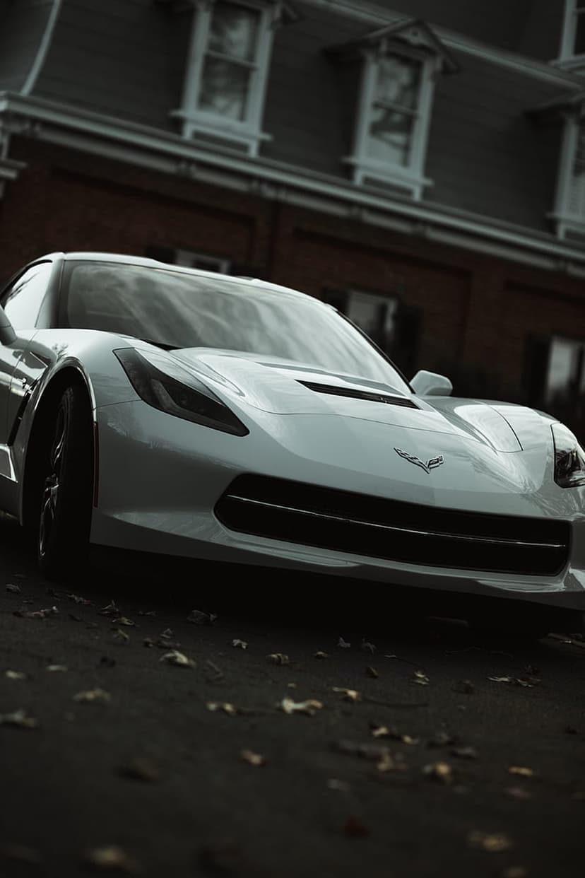 White Chevrolet Corvette C7 parked on dark asphalt with autumn leaves near a rustic red brick building facade.