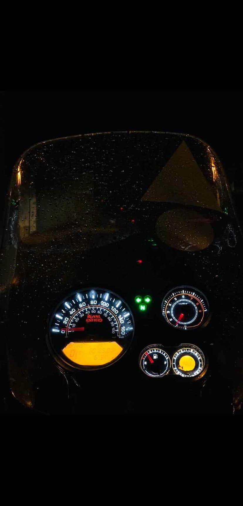 Royal Enfield Himalayan motorcycle dashboard glowing with amber light behind a rain-streaked windscreen at night.