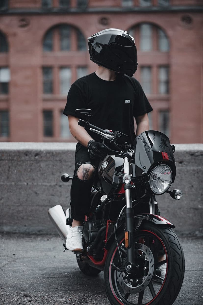 Biker in a black helmet and ripped jeans sits on a motorcycle with red accents against a brick wall.