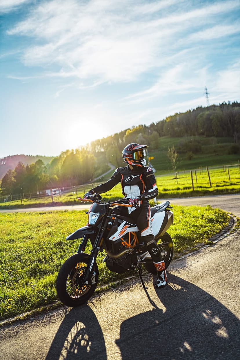 Biker on a black and orange supermoto sits in a grassy field during a vibrant golden hour sunset with trees.