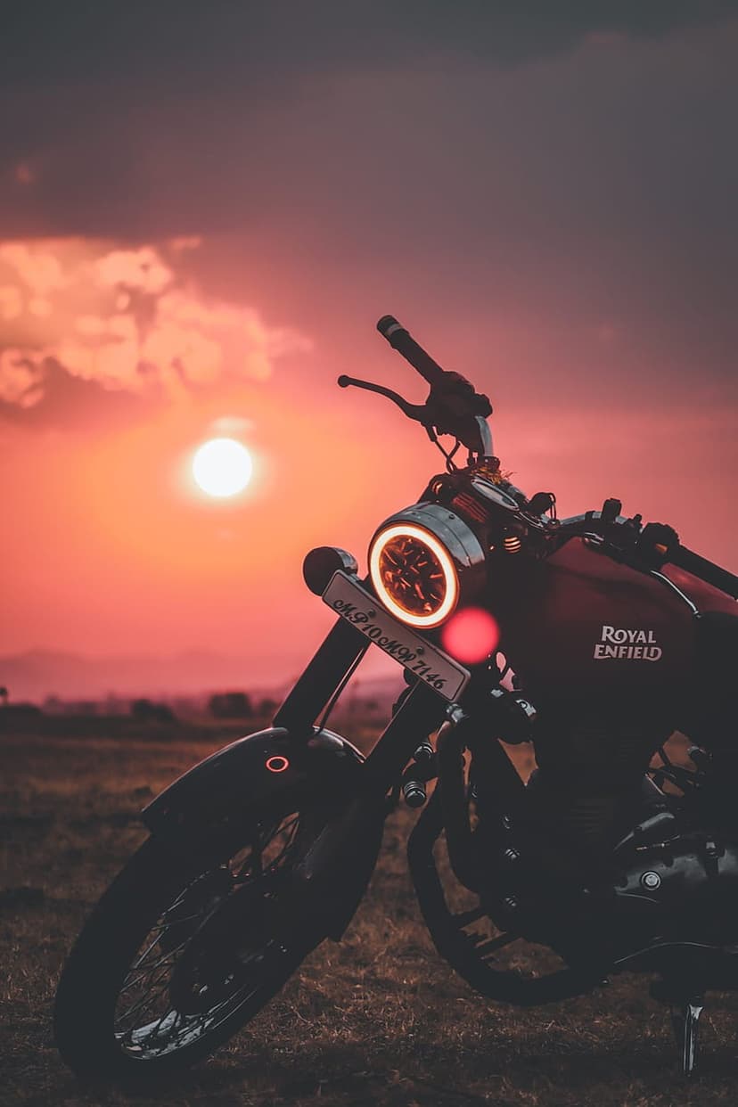 Royal Enfield motorcycle silhouetted against a fiery orange sunset with a glowing circular LED headlight.