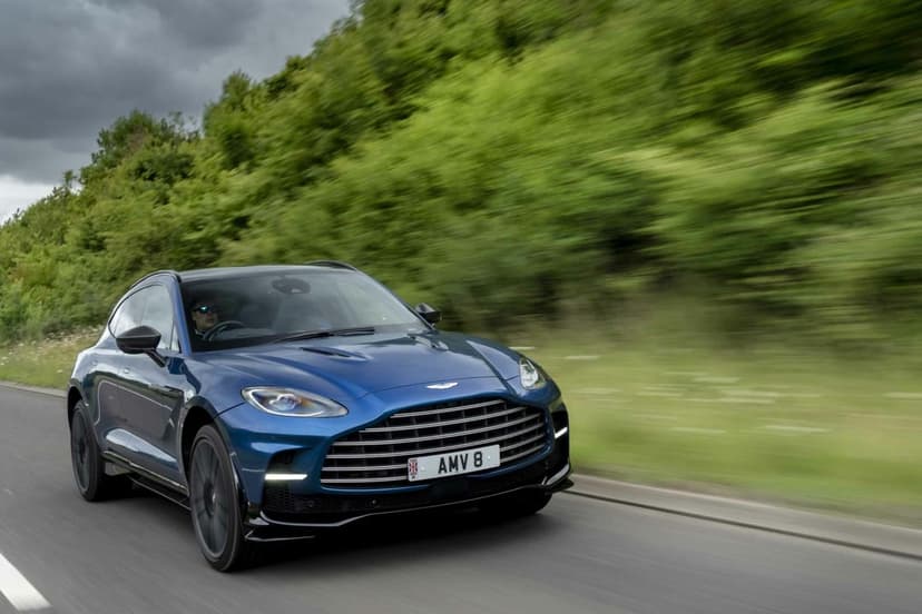 Blue Aston Martin DBX SUV driving fast on a road with motion-blurred green trees and an overcast sky.
