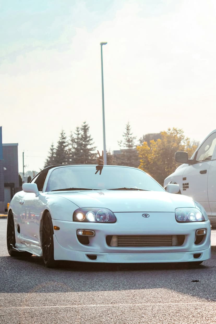 White Toyota Supra with glowing headlights parked on asphalt in a sunlit lot next to green trees and a truck.