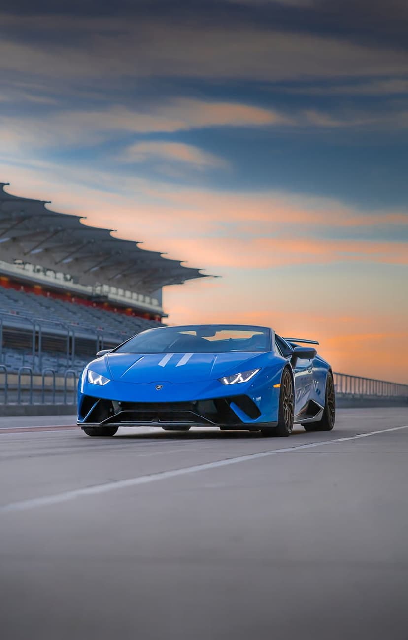 Blue Lamborghini Huracan Spyder parked on a racetrack asphalt under a vibrant orange and purple sunset sky.