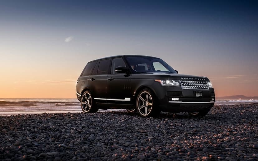 Black Range Rover SUV with HRE wheels parked on a pebble beach at sunset with ocean waves in the background.