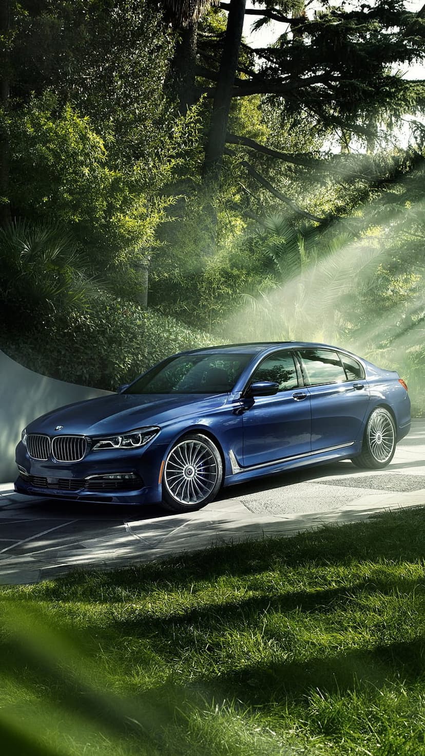 Blue luxury sedan parked on a stone pathway under sunlight filtering through green trees in a lush garden.