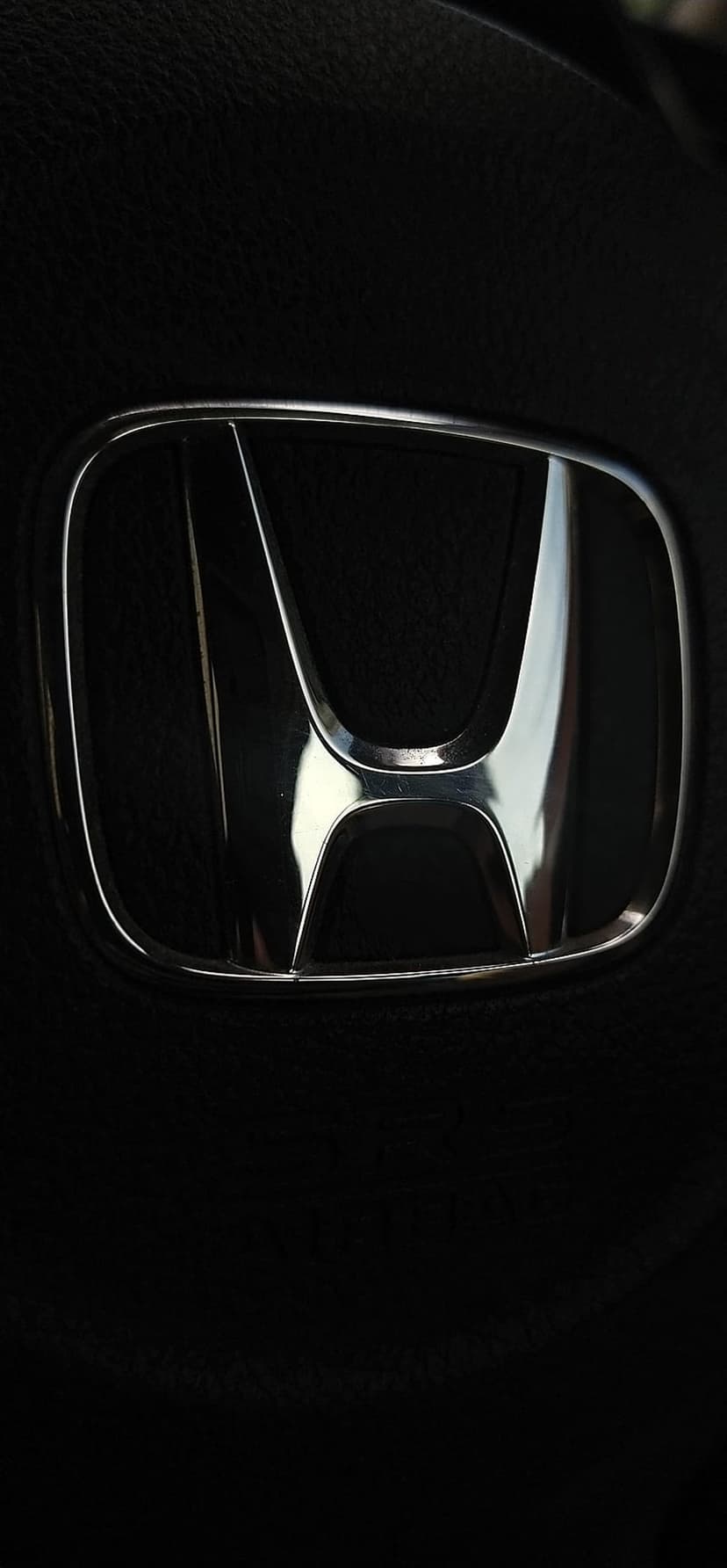 Honda logo badge in reflective chrome finish on dark textured leather upholstery with bright highlights.