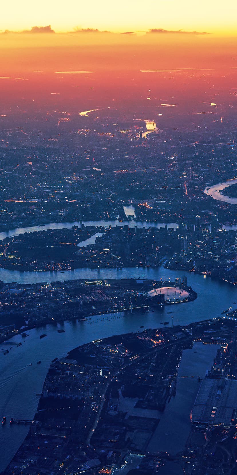 Aerial view of the River Thames snaking through glowing London buildings under a twilight sky at dusk.