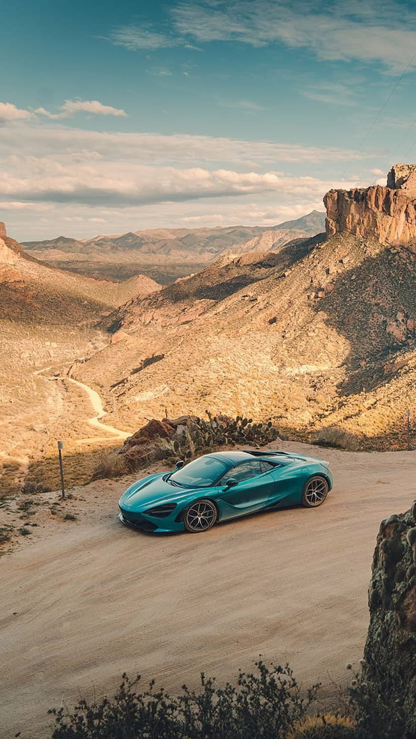 McLaren 720S in teal finish speeds along a dusty desert road beneath jagged mountains and a bright blue sky.