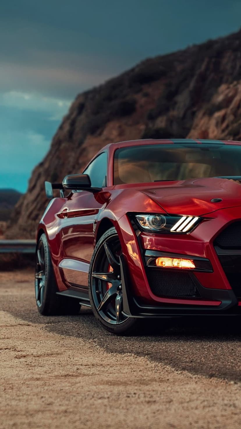 Red Ford Mustang GT500 parked on a gravel roadside with a backdrop of rugged mountains and a cloudy grey sky.