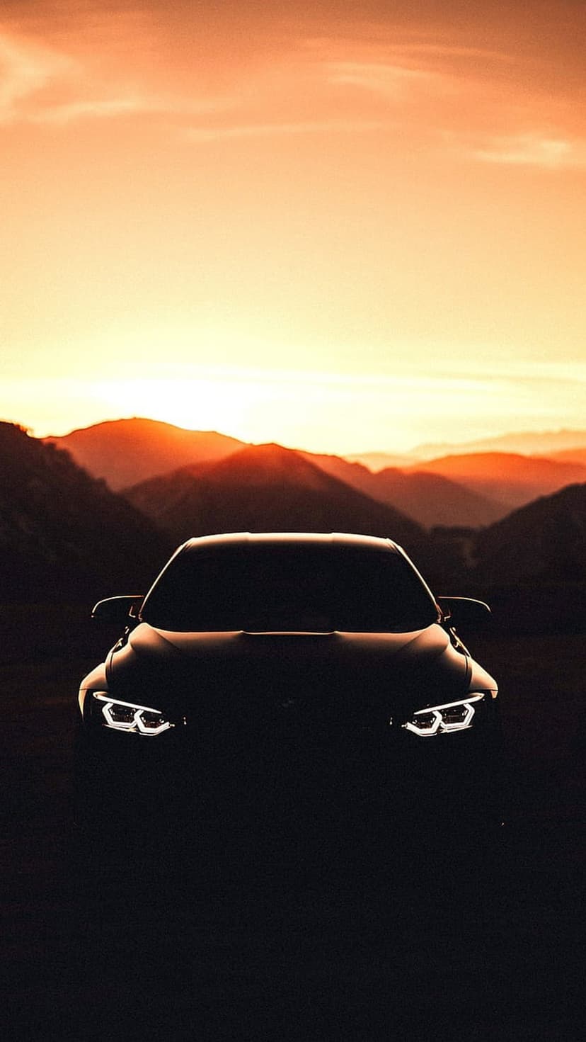 Glowing car headlights shine on a dark silhouette with layered mountains under an orange sunset sky.