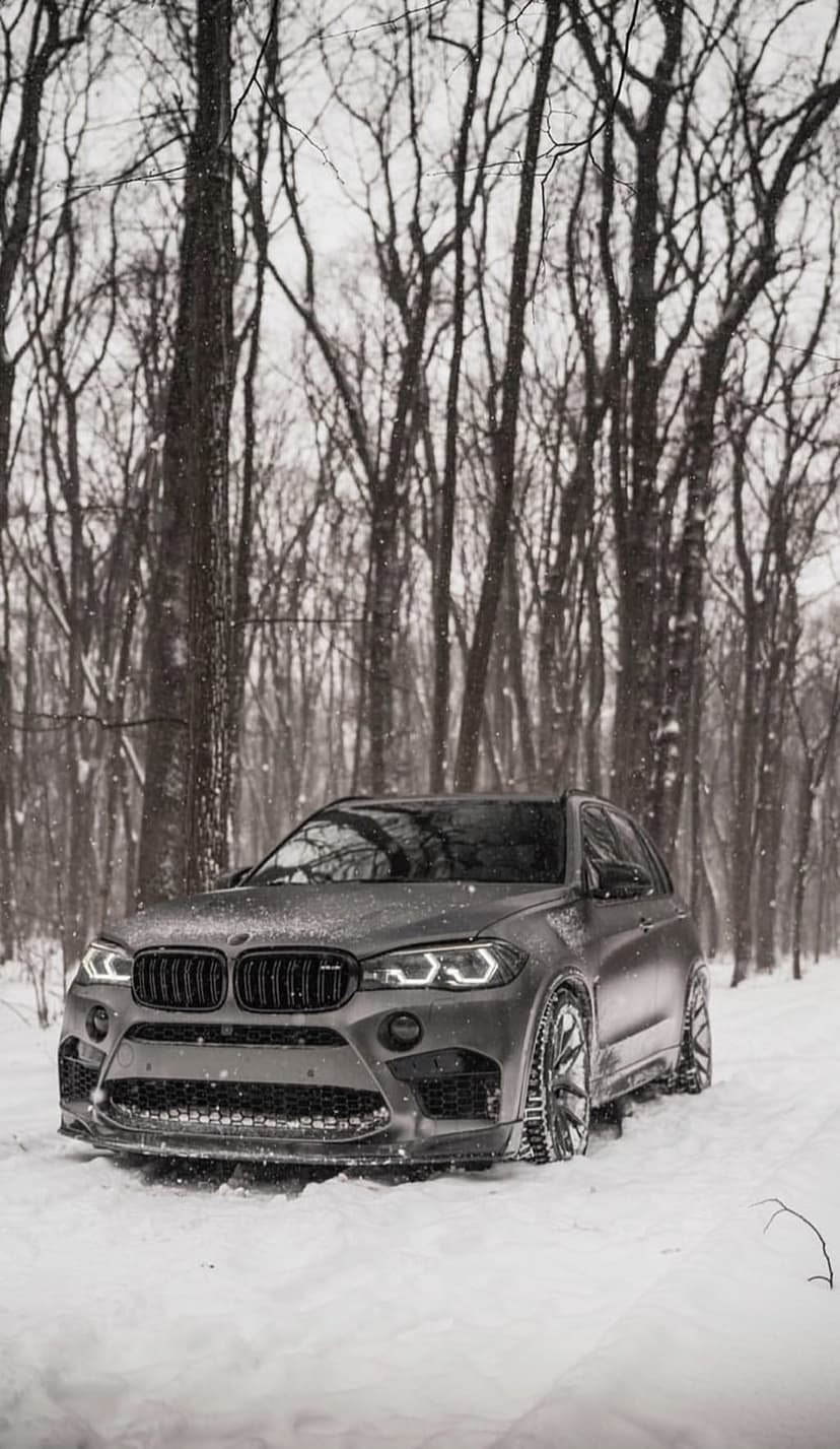 BMW X5 M SUV with glowing headlights stands on a snowy forest road amidst falling snow and bare winter trees.