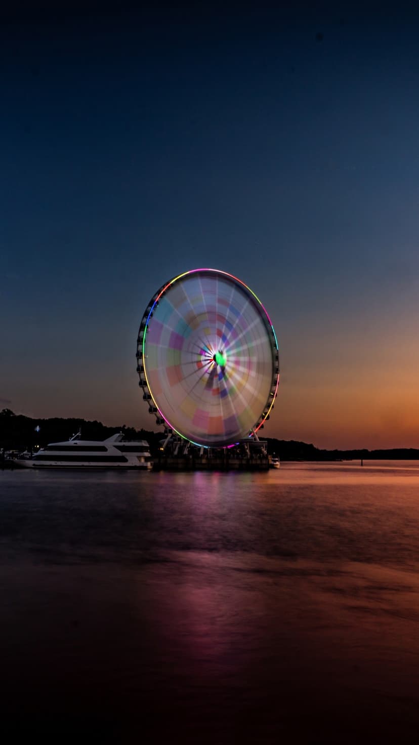 Illuminated Ferris wheel with colorful light streaks reflects on dark water beneath a gradient twilight sky.