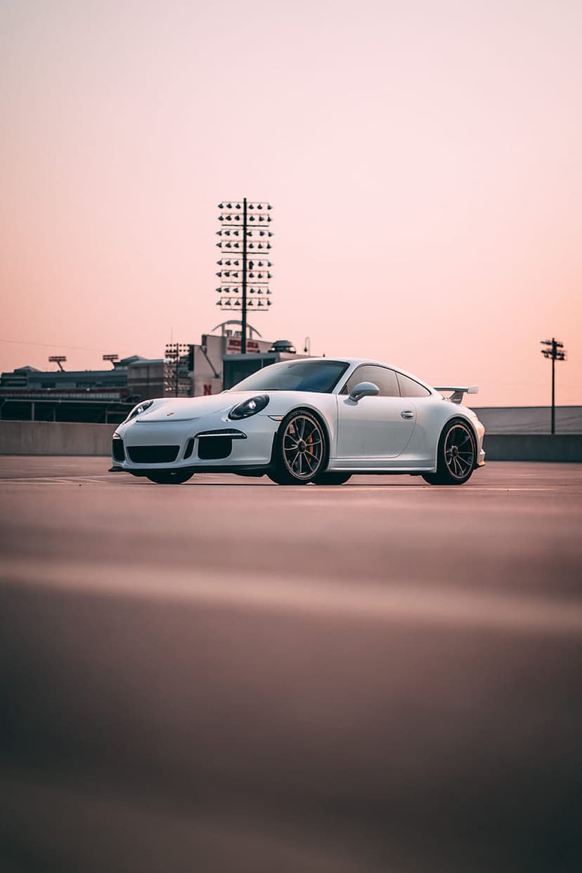 Porsche 911 GT3 in white parked on a concrete lot at sunset with stadium lights and urban structures behind.