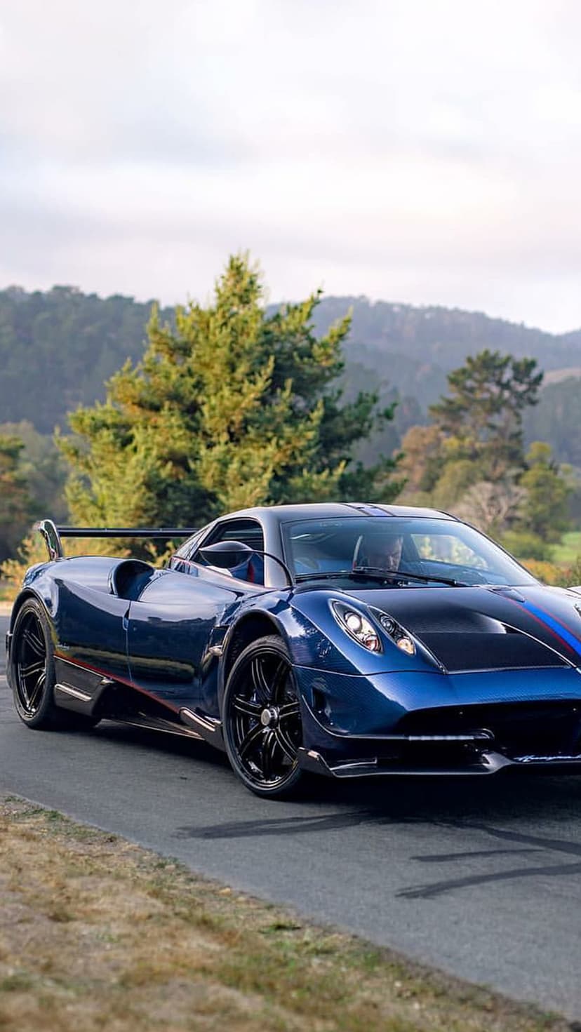 Blue Pagani Huayra BC Roadster driving on a scenic mountain road surrounded by green trees and lush hills.
