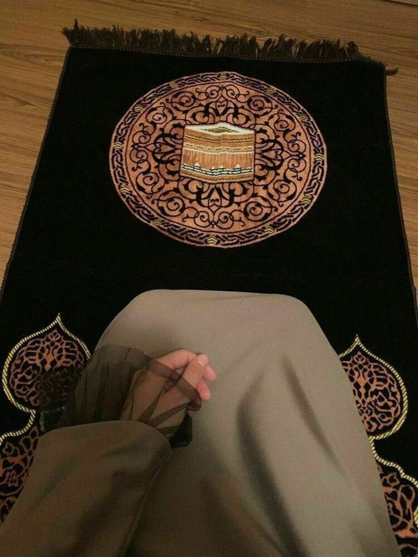 Woman in modest clothing kneels on a black prayer mat featuring a circular Kaaba embroidery and floral patterns.